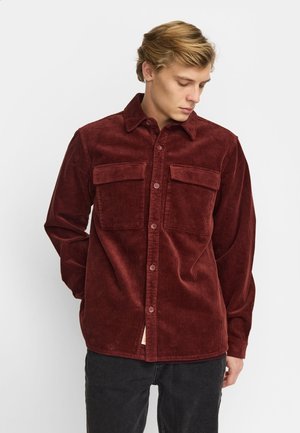 Man draagt een donkerrood corduroy overhemd met lange mouwen, dichtgeknoopt en met twee borstzakken, kijkt naar beneden, handen achter de rug.