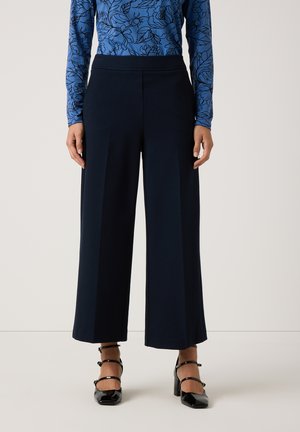 Marineblauwe wijde broek van zacht materiaal met een soepele textuur, voorzien van een hoge taille en geplooide voorkant. Draagt met zwarte bandjeshakken.