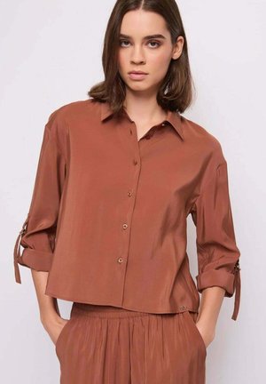 Donna con camicia marrone larga con bottoni e maniche arrotolate e gonna marrone abbinata, mani in tasca, su sfondo chiaro e semplice.