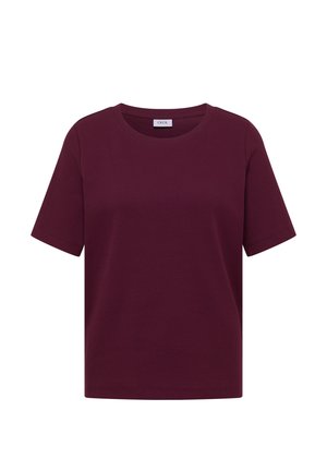 Frau mit langen braunen Haaren, die ein kurzärmeliges, geripptes, dunkelviolettes Shirt und hellblaue Jeans trägt, vom Kamera weg schauend.