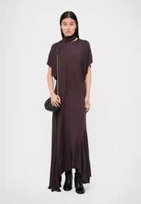 Long, flowing dress in deep purple fabric with a high neckline, featuring a draped design and short sleeves. Paired with a black handbag and boots.