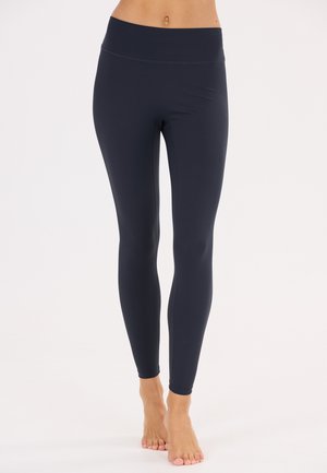 Kvinde iført højtaljede, tætsiddende sorte leggings, stående barfodet mod en ensfarvet hvid baggrund.