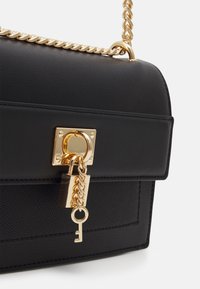 Černá kabelka z umělé kůže s texturovaným povrchem, vybavená zlatým řetízkovým popruhem, obdélníkovým tvarem a zlatým zámkem s klíčem.