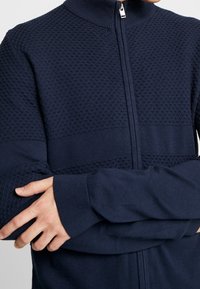 Marinblå stickad zip-up-tröja med ett texturerat diamantmönster på övre delen och släta nedre paneler, med hög krage och ribbade muddar.