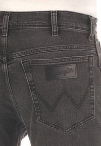 Los pantalones vaqueros de mezclilla negra cuentan con un bolsillo trasero con un diseño de "W" bordado y una etiqueta rectangular de cuero negro que muestra "Wrangler."
