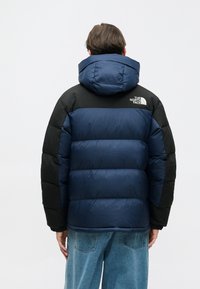 Tamnoplava i crna jakna na preklop s kapuljačom, s prošivenim uzorkom i detaljem logotipa na gornjem dijelu leđa. Dizajnirana za toplinu i udobnost.