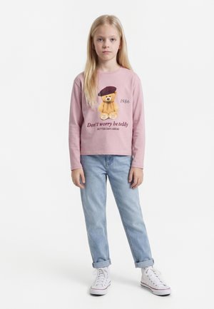 Νεαρή κοπέλα που φοράει ροζ μπλούζα με μακριά μανίκια, με γραφικό αρκουδάκι και κείμενο, ανοιχτό μπλε τζιν και λευκά αθλητικά παπούτσια, σε λευκό φόντο.