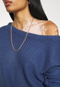 Pull bleu tricoté avec un design asymétrique dévoilant une épaule, en tissu texturé. Un collier chaîne en or pend sur l'encolure.