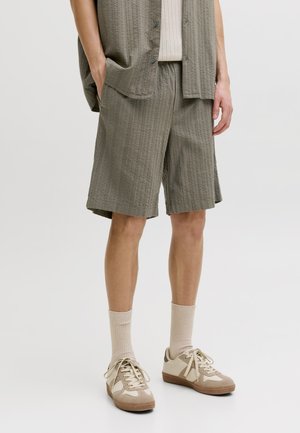 Person iført olivengrønne teksturerede knælange shorts, matchende åben skjorte, beige ribstrikkede sokker og beige sneakers med brune såler.