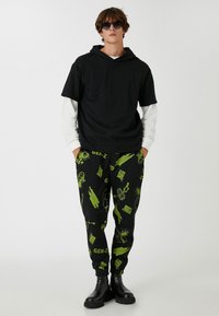 Kortärmad svart hoodie över en långärmad vit skjorta, i kombination med svarta joggers med gröna grafik. Svarta ankelskor fulländar looken.