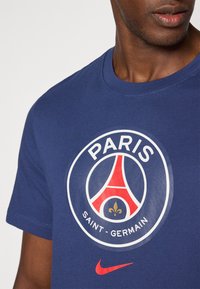Marineblå bomulds T-shirt med et stort rundt Paris Saint-Germain-logo i hvid, rød og guld. Inkluderer en rød Nike-swoosh accent.