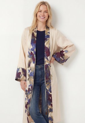 Femme souriante portant un long kimono beige avec une bordure florale violette et bleue, sur un haut bleu marine et un jean bleu, debout avec une main sur la hanche.
