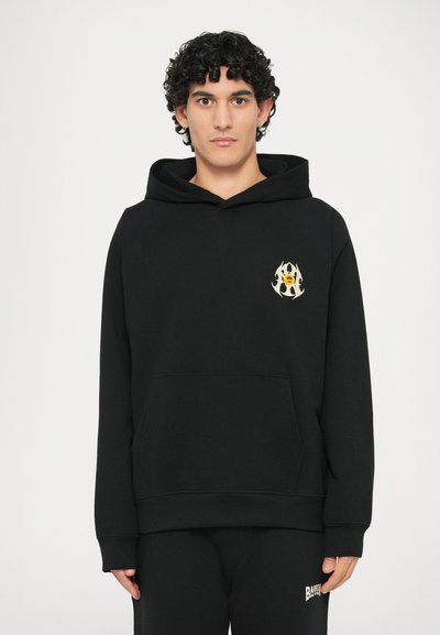 Čierna mikina s kapucňou a prednou vreckom, s žltým a bielym plameňom na hrudi. Vyrobená z mäkkej, štruktúrovanej tkaniny.
