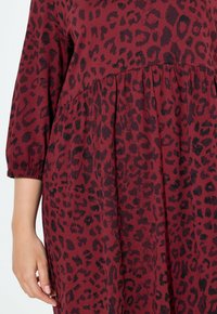 Burgundy-mekko, jossa on musta leopardikuvio. Kankaassa on pehmeä rakenne, ja siinä on tyköistuva vyötärö ja kyynärpituiseen asti ulottuvat hihat.