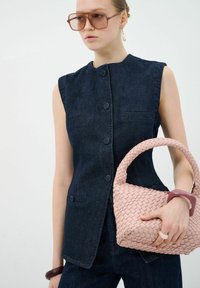 Denim vest met een hoge kraag, donkerblauwe kleur, knopen en zijzakken. Model houdt een textuur roze geweven handtas vast.