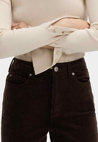 Beige gerippter Strickoberteil mit Knöpfen an den Ärmeln, kombiniert mit hochtaillierten, dunkelbraunen Cordhosen mit frontalen Taschen und Metallakzenten.