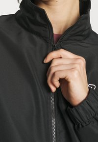 Svart zip-up jacka med ståkrage och elastiska muddar. Har en texturerad tygkvalitet och en liten logodetalj på vänster bröst.