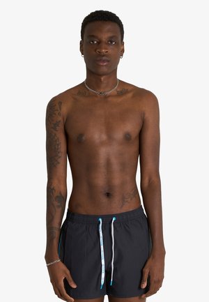Giovane uomo con capelli corti e ricci, tatuaggi sul braccio sinistro e sul petto, indossa pantaloncini da bagno neri con cordini bianchi e blu, in piedi su sfondo bianco.