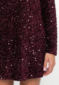 Burgundské šaty s flitrami a dlhými rukávmi, s textúrovaným povrchom pokrytýmLesklými, reflexnými flitrami, ktoré zachytávajú svetlo.