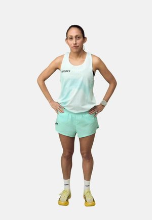 Atlet stående med hænderne på hoften iført en hvid Brooks tanktop, mintgrønne shorts, hvide sokker og gule løbesko.
