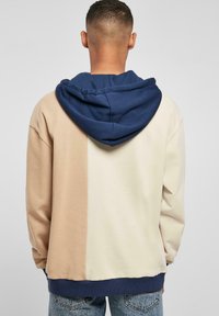 Hoodie v blokových farbách v béžovej, krémovej a námorníckej modrej. Mäkkučká tkanina, s veľkou kapucňou a ribovanými manžetami. Voľný strih. Zadný pohľad ukazuje detaily švov.