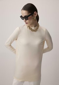 Hosszú ujjú, bézs felső magas gallérral, sima textúrával. Arany láncnyaklánccal és fekete, négyzet alakú napszemüveggel viselve.