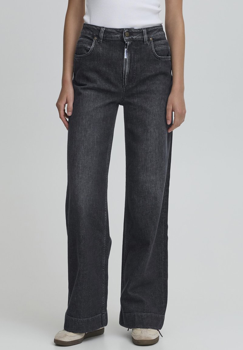 Plati kojinės juodos džinsinės kelnės su lygiu paviršiumi, turinčios priekinės kišenes, mygtuką ir užtrauktuką, bei šiek tiek išblukusį paviršių.