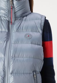 Gilet matelassé bleu clair avec un col haut, fermeture éclair et patch logo sur la poitrine. Texture lisse et brillante avec un design matelassé.