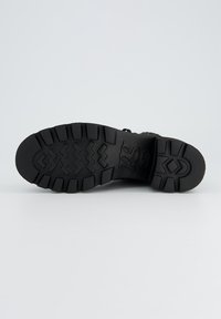 Semelle de chaussure en caoutchouc noir avec un motif de bande de roulement en zigzag, présentant une surface texturée et un logo de marque en relief au centre.