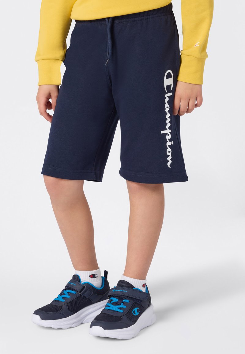 Enfant portant un short Champion bleu marine avec un logo blanc sur le côté, des chaussures de sport bleu marine et bleu vif, des chaussettes blanches, et un t-shirt à manches longues jaune.