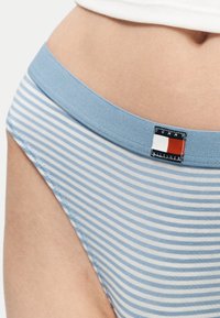 Μπλε και άσπρο ριγέ εσώρουχο με ανοιχτό μπλε ζωνάρι στη μέση που φέρει ένα ετικέτα με το λογότυπο της Tommy Hilfiger στο ισχίο ενός ατόμου.