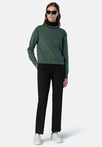 Maglione verde con collo alto, sottoshirt nero, pantaloni neri su misura e scarpe da ginnastica bianche; presenta polsi a coste e linee pulite.