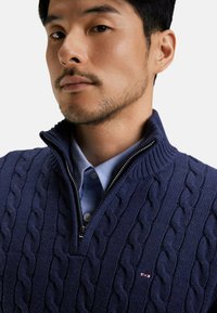 Pull en maille bleu marine avec col montant, fermeture éclair et une chemise à col bleu clair en dessous. Petit logo sur la poitrine gauche.