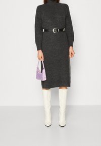 Persona che indossa un vestito a coste grigio scuro con una cintura nera, mentre tiene una piccola borsa lavanda e indossa stivali bianchi alti fino al ginocchio.