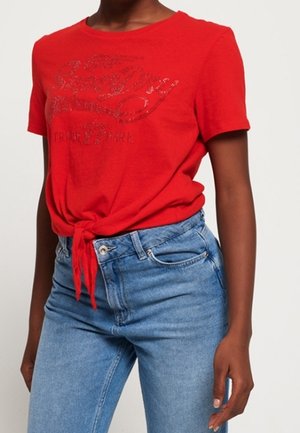 Red short-sleeve T-shirt with front knot and glittery "SuperDry" logo paired with high-waisted light blue jeans on a standing person.