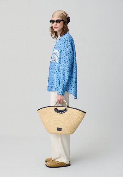 Camicia a fantasia blu, pantaloni bianchi, borsa di paglia con accenti neri e hardware dorato, occhiali da sole e un foulard marrone chiaro.
