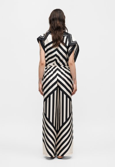 Γυναίκα που στέκεται με την πλάτη, φοράει μαύρο-άσπρο ριγέ παντελόνι με φαρδιές μπατζάκια και ταιριαστό τοπ με κοντά μανίκια και βολάν στους ώμους.