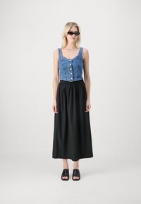 Denim apgrieztais tops ar pogām, saskaņots ar melnu maksisvārku; modele valkā melnas saulesbrilles un atvērtas melnas kurpes.