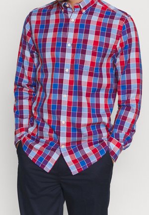 Homme portant une chemise à manches longues boutonnée avec un motif à carreaux rouge, bleu et violet, et un pantalon foncé, mains dans les poches.