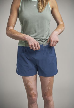 Person mit Vitiligo, die den Bund blauer Sportshorts anpasst, trägt ein ärmelloses hellgrünes Oberteil vor neutralem Hintergrund.