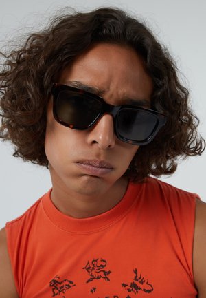 Tortoiseshell zonnebril met donkere lenzen. Dikke montuur met een vierkante vorm. Onderwerp draagt een oranje mouwloos shirt met een grafische print.