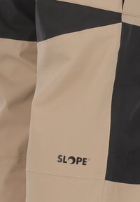 Beige och svarta skidbyxor med en jämn textur. Har "SLOPE"-logotypen i svart på nedre vänstra delen. Distinkt färgblockeringsdesign.