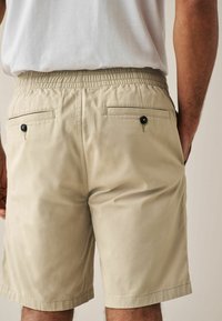 Pantaloni corti beige con vita elastica, due tasche posteriori con bottoni neri, texture liscia e vestibilità sartoriale.
