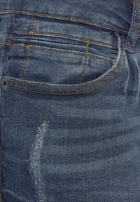 Close-up van de zak van een blauwe spijkerbroek met oranje stiksels en een vervaagde versleten plek op het bovenbeen.