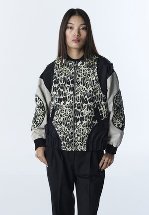 Giovane donna con capelli lunghi che indossa una giacca con zip con pannelli a stampa leopardata e maniche nere e beige, in piedi davanti a uno sfondo semplice.