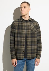 Camisa de franela a cuadros verdes y negros con cierre de botones, mangas largas y un bolsillo en el pecho, llevada sobre una camiseta negra.