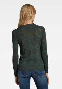 Suéter de punto verde oscuro con un patrón de diamantes, superficie texturizada y puños y dobladillo acanalados. La vista trasera muestra el detalle de la marca.