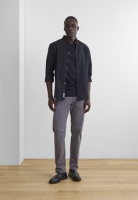 Navy button-up shirt over a dark t-shirt, gray pants, and black shoes. Features a relaxed fit with rolled sleeves and a label detail.