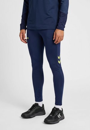 Osoba nosząca granatowe sportowe legginsy i pasującą bluzkę, w połączeniu z czarnymi butami sportowymi i białymi skarpetkami, stojąca na szarej podłodze.