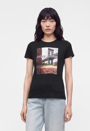 Femme aux longs cheveux noirs porte un t-shirt noir avec une photo du pont de Brooklyn et un jean bleu clair, debout devant un fond blanc uni.